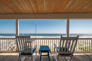 A wooden deck with two white chairs and a small table, overlooking a beach with the ocean and a clear sky. - Beach Barracks: Simple, Oceanfront Home with Large Covered Porch and Dune-top De (Oak Island)