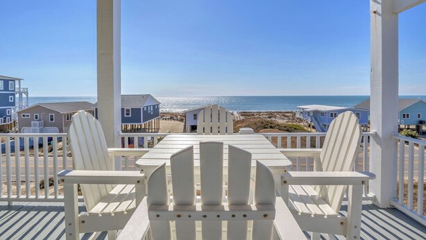 Um deck branco com mesa e cadeiras, com vista para uma praia, casas e o oceano.