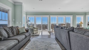 A spacious living room with a large sectional sofa, a small round table, and a view through large glass doors to an outdoor balcony. - Turtle Soup: Oceanfront home in East Beach with plenty of ocean leading windows (Oak Island)