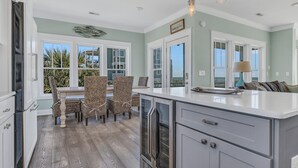 A kitchen with a large island, white cabinetry, and a dining area with wicker chairs. - Turtle Soup: Oceanfront home in East Beach with plenty of ocean leading windows (Oak Island)