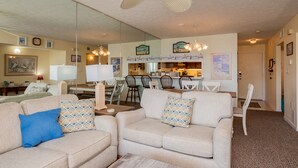 A living room with a beige sofa, a coffee table, and a ceiling fan. There is a kitchen area with a dining table and chairs visible through a glass partition. - A Pelican's Paradise: Condo with Community Pool and Ocean Views (Caswell Beach)