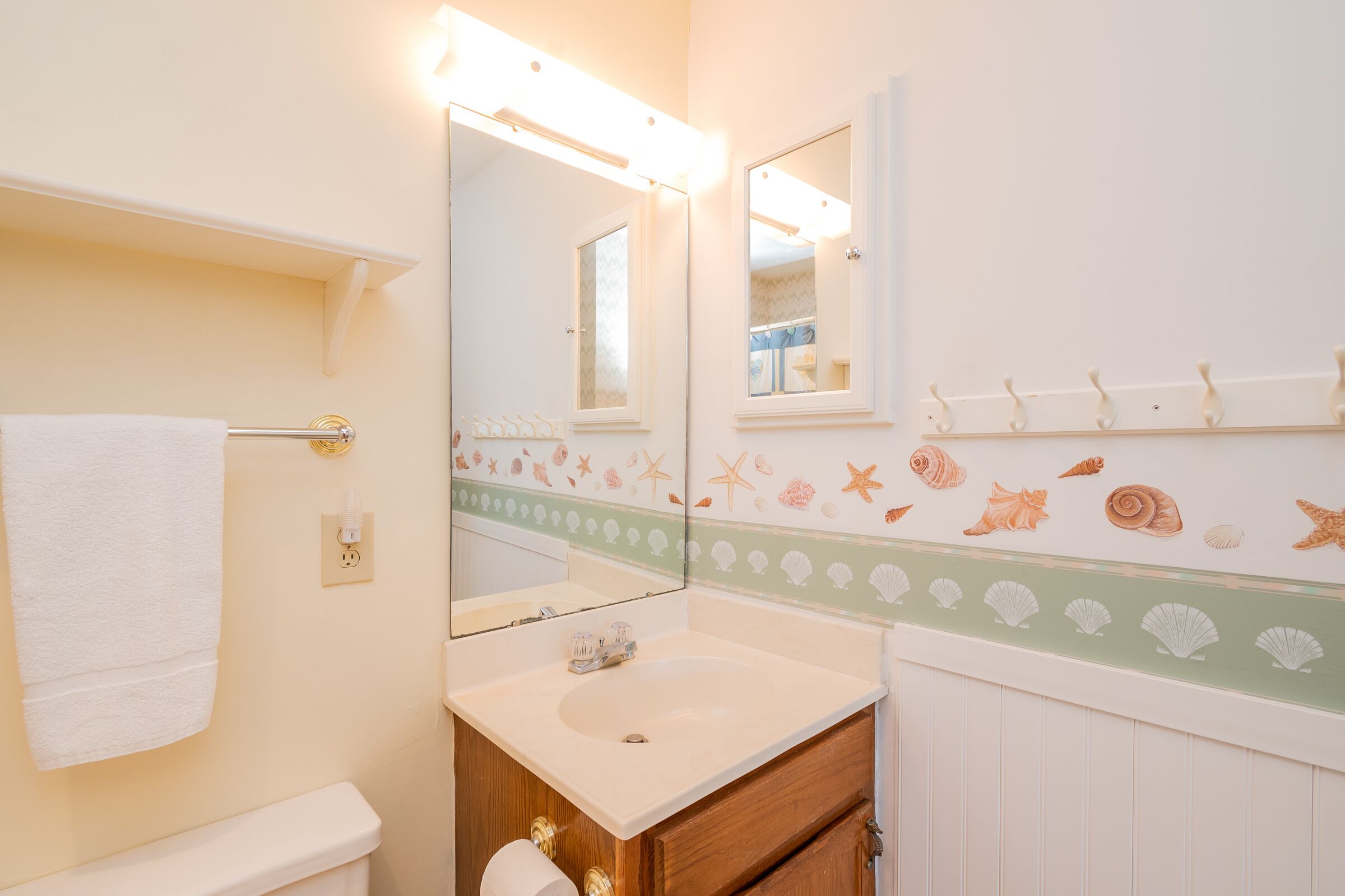 Een badkamer met een witte wasbak, een spiegel, een handdoekhouder en een decoratieve wand met schelpen en zeesterren.