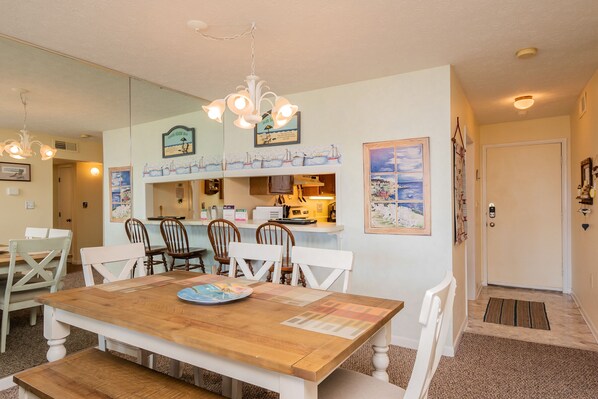 A dining area with a wooden table, white chairs, and a chandelier. There is a kitchen area in the background with a counter and various items on it. - A Pelican's Paradise: Condo with Community Pool and Ocean Views (Caswell Beach)