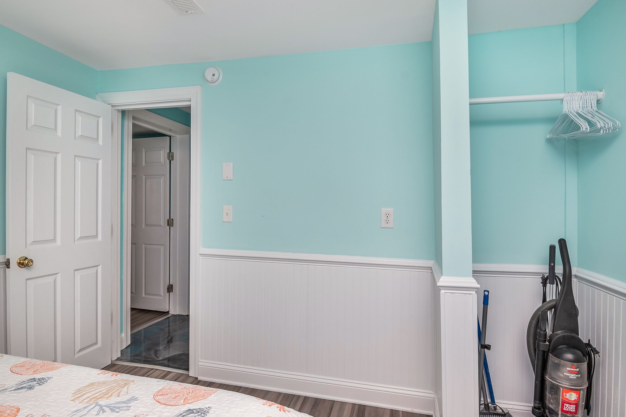A room with teal walls, white wainscoting, a door leading to another room, a vacuum cleaner, and a clothes rack.