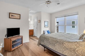 Ein Schlafzimmer mit einem Bett, einem Fernseher auf einem Holzschrank, einem Deckenventilator und einem großen Fenster mit Jalousien.
