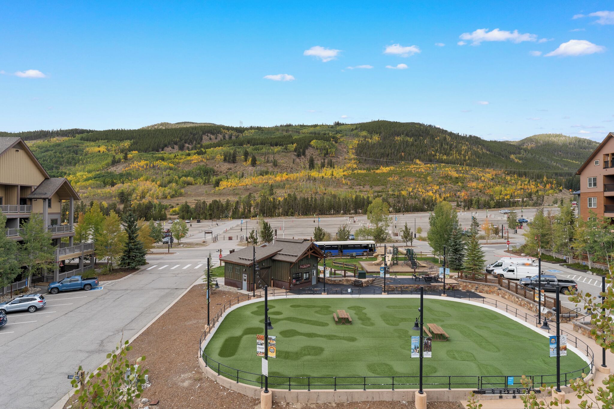 A hotel with a parking lot, a small building with a sign, and a view of a hilly landscape with autumn-colored trees.
