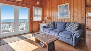 A wooden cabin interior with a gray sofa, a wooden coffee table, and a view of the ocean through glass doors.
