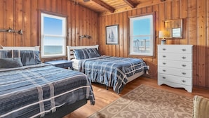A wooden cabin bedroom with two beds, a dresser, a ceiling fan, and large windows.