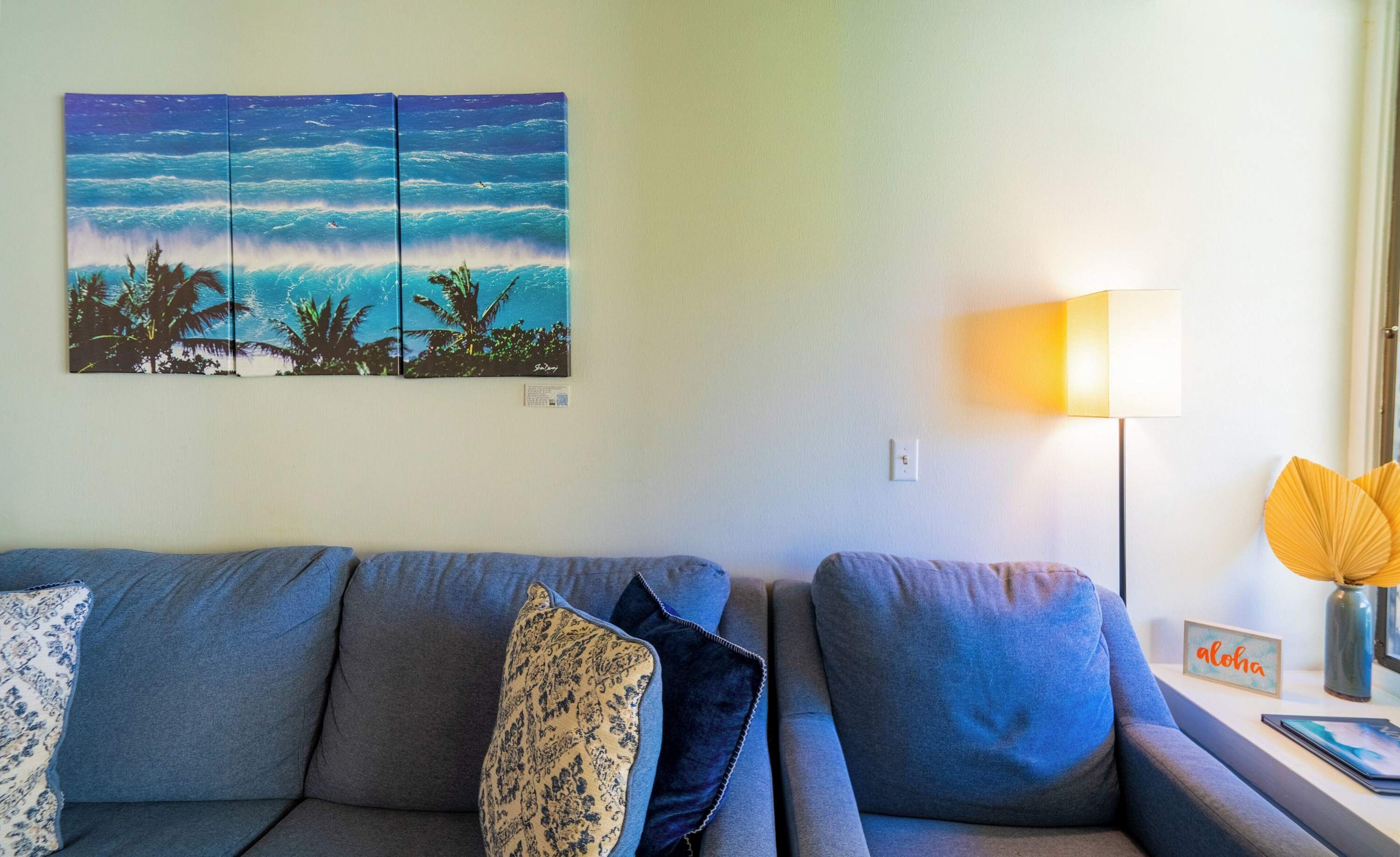 A living room with a grey sofa, a wall-mounted painting of a tropical scene, a floor lamp, and a small table with a vase and a sign.