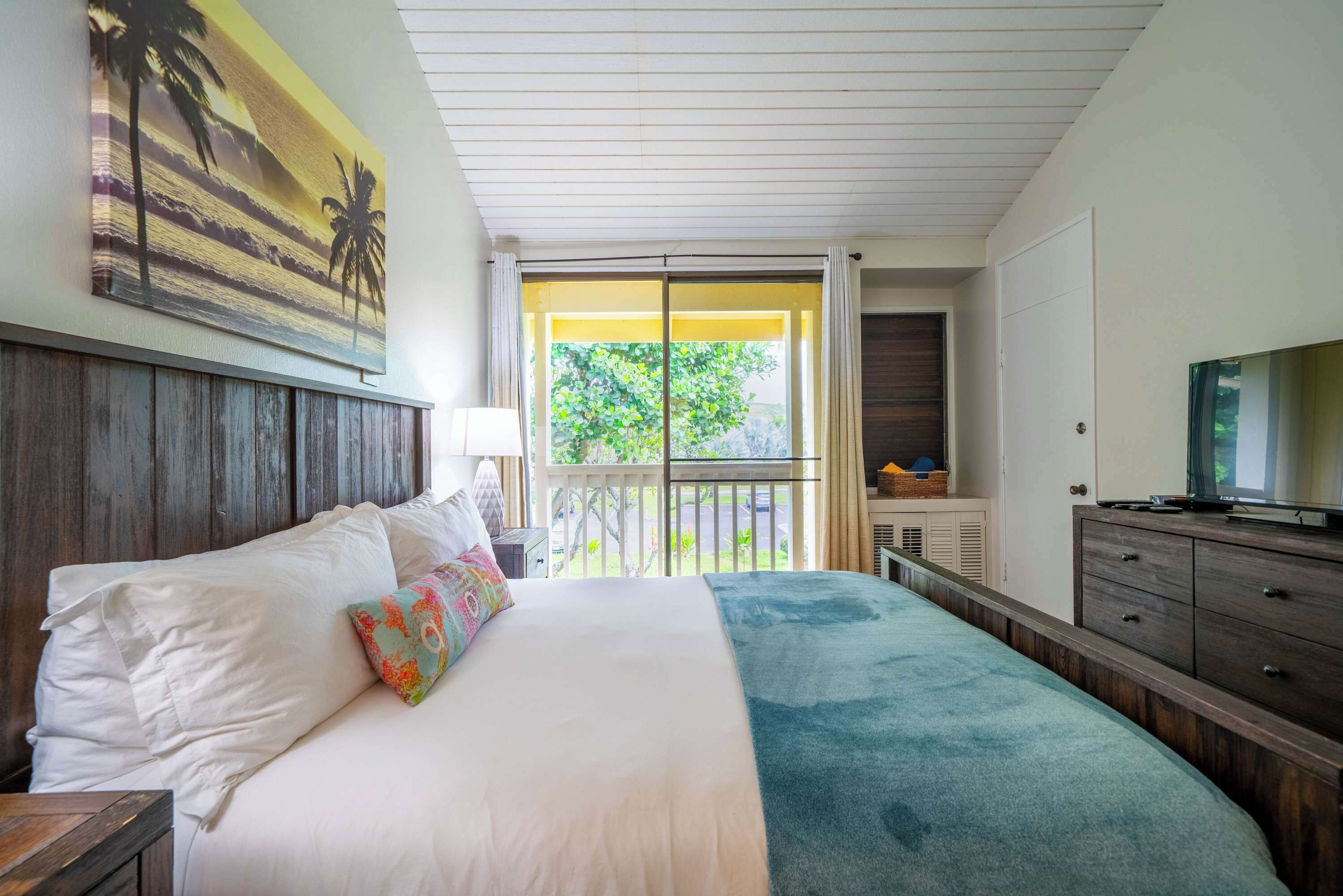 Un dormitorio con cama, televisor, cómoda y balcón con vistas a zonas verdes.
