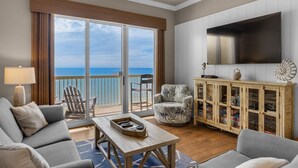 A spacious living room with a large sliding glass door leading to a balcony with a view of the ocean. There is a flat-screen TV mounted on the wall, a wooden cabinet with glass doors, and a comfortable seating area with a sofa and armchairs.