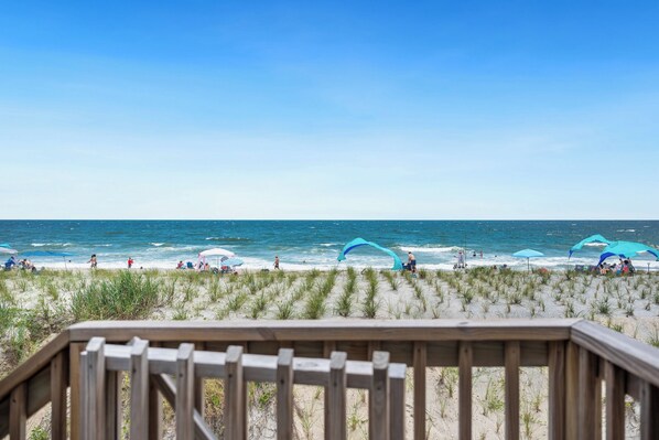 A beach with a wooden deck in the foreground, a sandy beach with grass, and the ocean in the background.