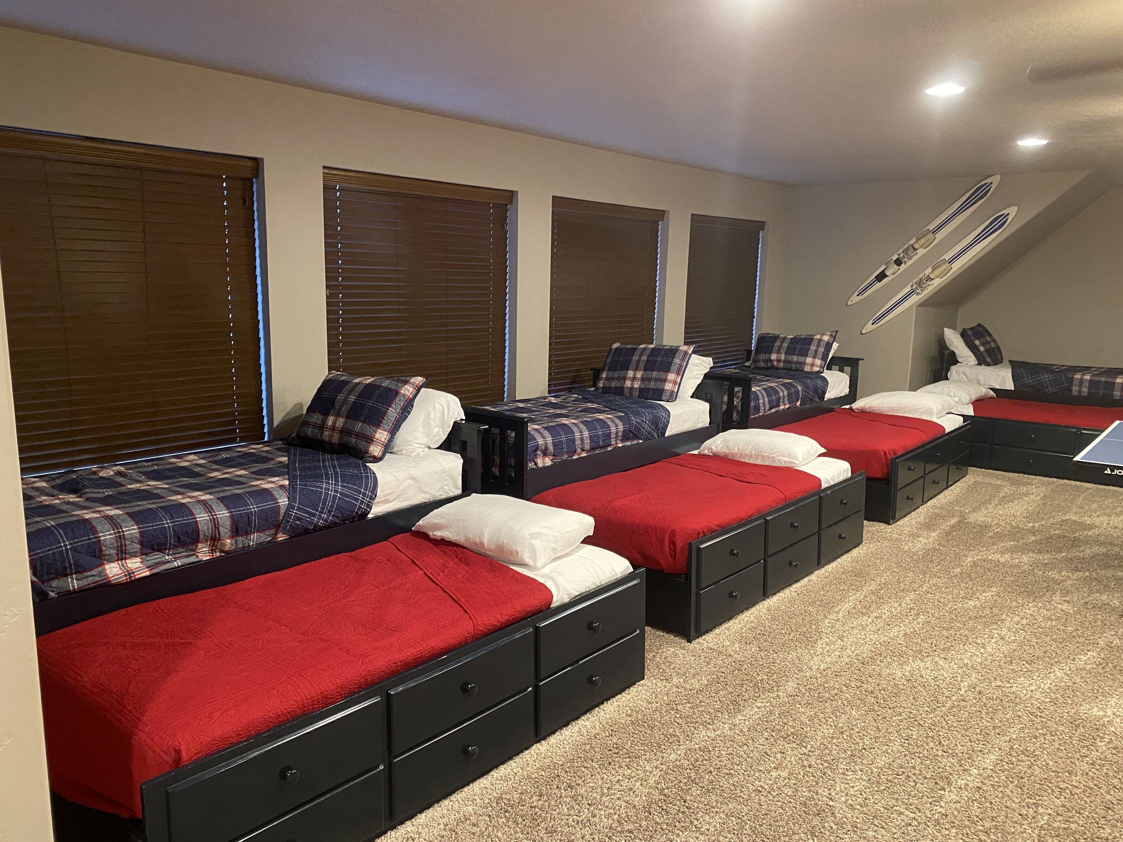 Une chambre avec quatre lits alignés, chacun recouvert d’une couverture à carreaux rouges et bleus, et un ventilateur de plafond au-dessus.