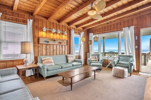 A cozy room with wooden walls, a sofa, a coffee table, and a view through large windows. - Boys Oh Boy: Oceanfront House in West Beach (Oak Island)