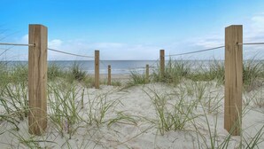 Une clôture de plage en bois, agrémentée d’une barrière en corde, mène à une plage de sable où l’on aperçoit de l’herbe au premier plan.