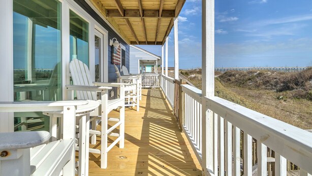 Une terrasse en bois avec des rampes blanches, des sièges d’extérieur et une vue sur la plage et la jetée.