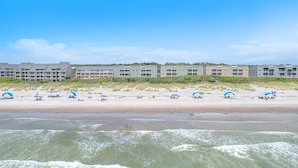 A beachfront hotel with multiple floors, a sandy beach with umbrellas, and a view of the ocean.