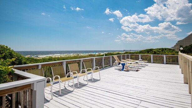 A wooden deck with white chairs and a blue chair, overlooking a beach and ocean.