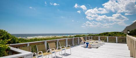 A wooden deck with white chairs and a blue chair, overlooking a beach and ocean.