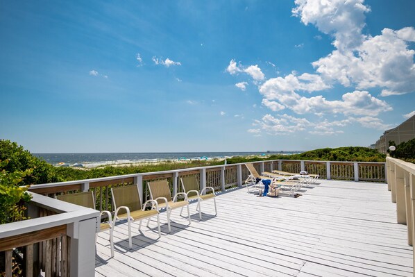 Terraza de madera con sillas blancas y una silla azul, con vista a una playa y al océano.