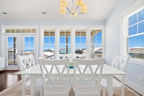A dining area with a white table and chairs, large windows with a view, and a chandelier. - Kiki Hut: Swimming Pool, Great Ocean Views, Dog-Friendly (Oak Island)