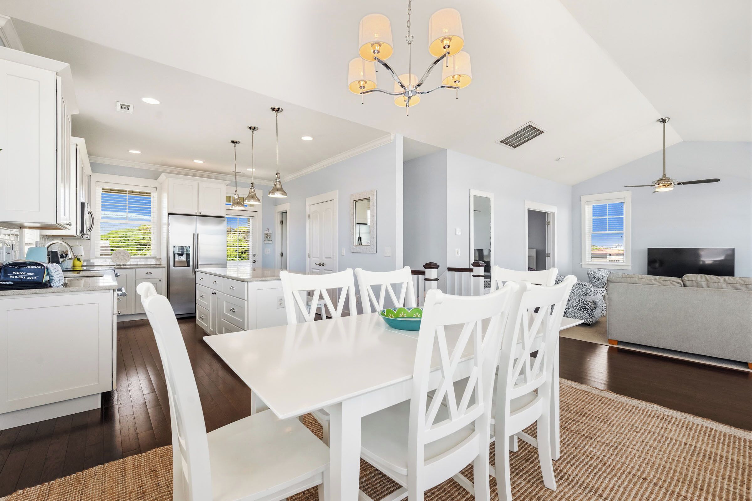 A modern kitchen with white cabinets, a dining table with white chairs, a chandelier, and a living area with a gray sofa.
