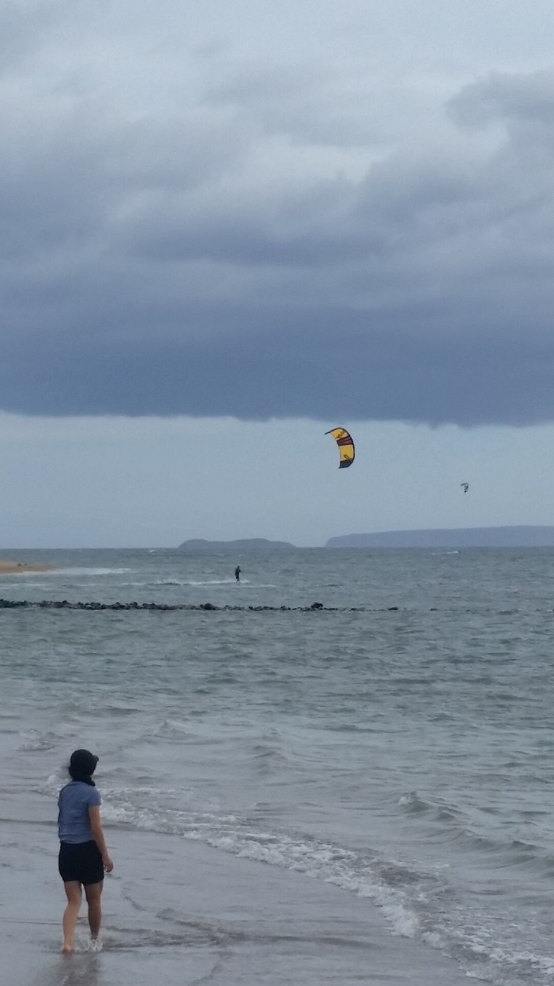 A beach with a person walking towards the water, a kite surfer in the distance, and a cloudy sky.