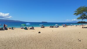 Una platja amb gent relaxant-se sota ombrel·les, un cel clar i una illa a la distància.