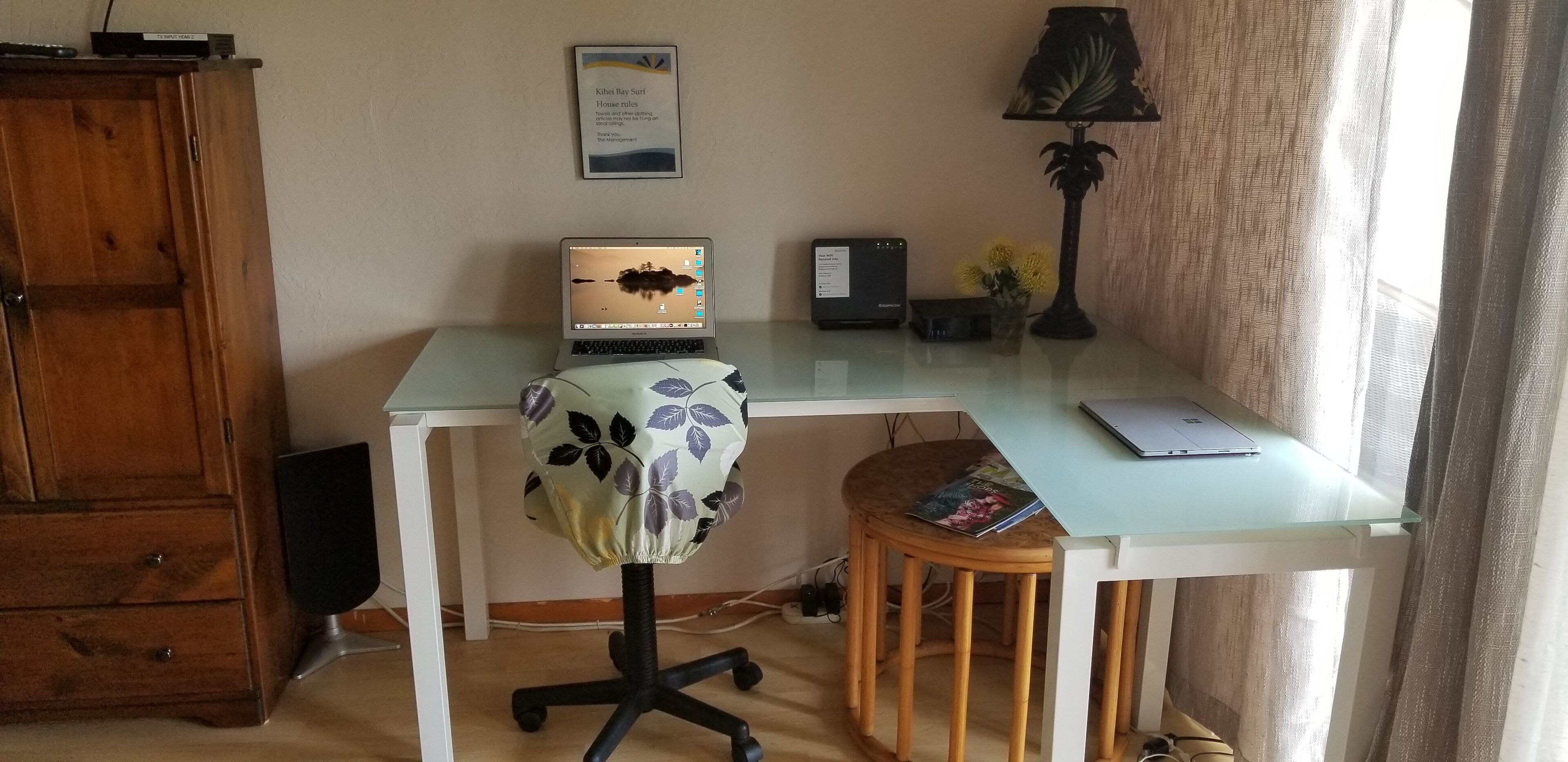 A home office setup with a laptop on a glass desk, a wooden chair, a lamp, and a framed certificate on the wall.