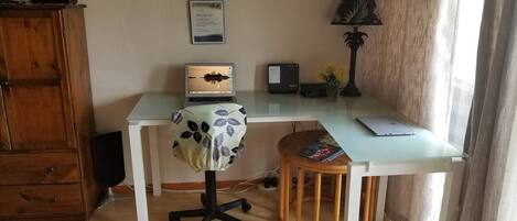A home office setup with a laptop on a glass desk, a wooden chair, a lamp, and a framed certificate on the wall.
