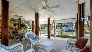 An outdoor patio area with wicker furniture, a ceiling fan, and a view of a water body and buildings in the background. - Wine Down Large Corner Canal Lot - Sleeps 8 (Jamaica Beach)