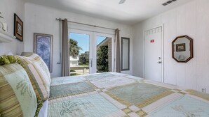 A bedroom with a large bed, a ceiling fan, and a view through the open door. - Wine Down Large Corner Canal Lot - Sleeps 8 (Jamaica Beach)