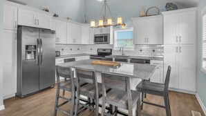Una cocina moderna con armarios blancos, un refrigerador de acero inoxidable, una isla central con taburetes altos y una lámpara de araña.