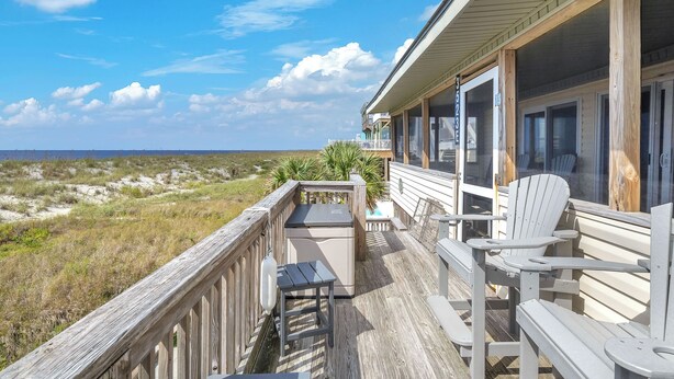 Una terraza de madera con vistas a la playa, un área de estar cubierta con sillas y una mesa pequeña.