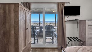 Una habitación con puerta corrediza de vidrio que da a una terraza exterior con sillas y una mesa, un televisor de pantalla plana en la pared y un armario de madera.