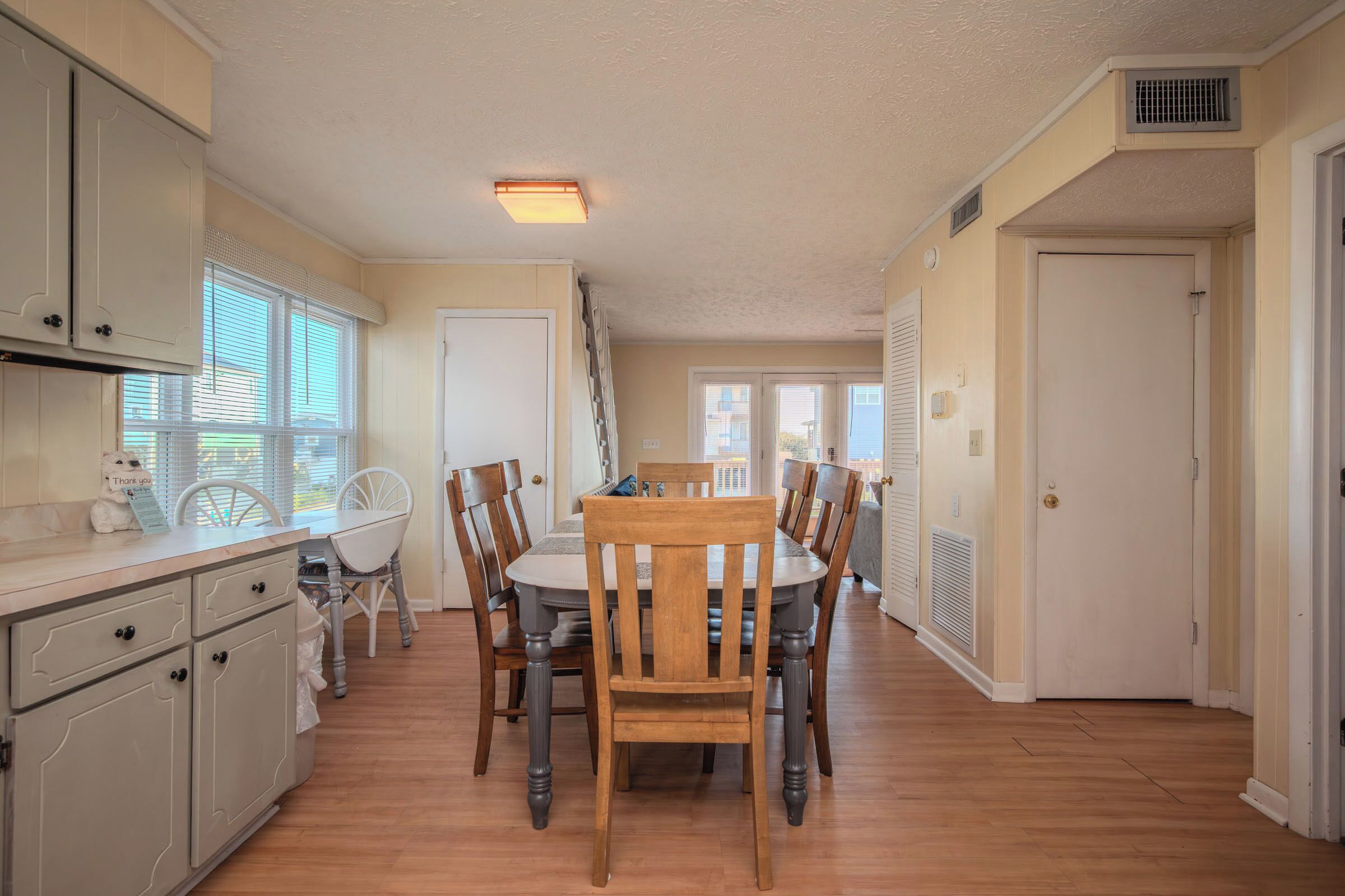 A dining area with a round table and six chairs, a kitchen with white cabinets, and a view of the outside through a window.