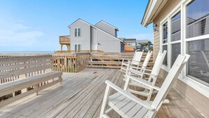 Una terraza de madera con sillas blancas y un banco de madera, con vistas a una playa y una casa de dos pisos con balcón.