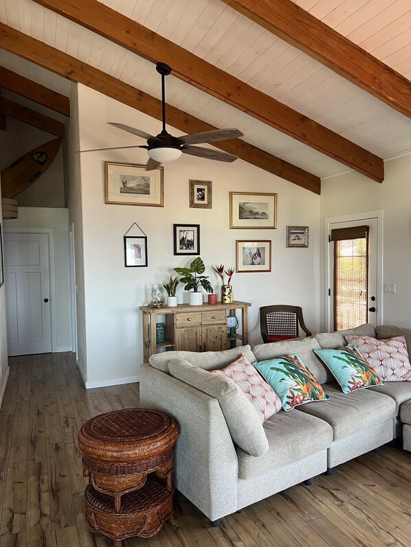 Ein gemütliches Wohnzimmer mit Holzboden, Deckenventilator, grauem Sofa mit gemusterten Kissen, einem Korbstuhl und einer Holzkabine mit Dekoration.