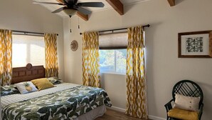 Une chambre à coucher avec un lit, une chaise, un ventilateur de plafond et une fenêtre avec des rideaux.