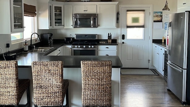 Une cuisine moderne avec des meubles blancs, un îlot central doté de chaises en osier et un ventilateur de plafond.