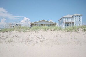 A beachfront property with a main house and a larger building, surrounded by sand and sparse vegetation.