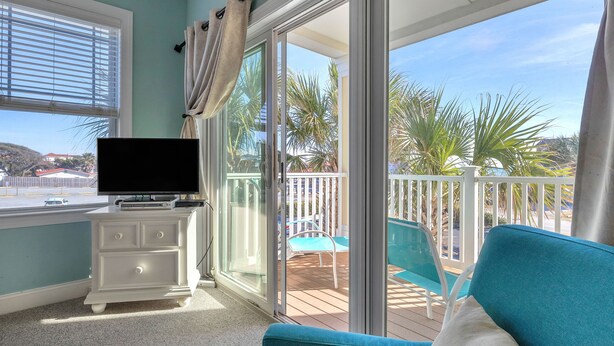 A balcony with a white dresser, a teal sofa, and a television on the dresser. - Beats Working: Condo with Community Pool Near OKI Pier and Restaurants (Oak Island)
