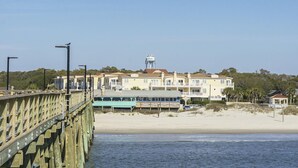 A beachfront property with a wooden pier, a large multi-story building, and a beach in the background. - Beats Working: Condo with Community Pool Near OKI Pier and Restaurants (Oak Island)