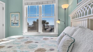A bedroom with a large window, a bed with a patterned quilt, a wall-mounted lamp, and a framed picture of a sailboat. - Beats Working: Condo with Community Pool Near OKI Pier and Restaurants (Oak Island)