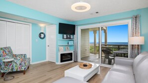 Ein Wohnzimmer mit Sofa, Couchtisch, Fernseher und Kamin. Ein Balkon mit Tisch und Stühlen ist ebenfalls vorhanden.