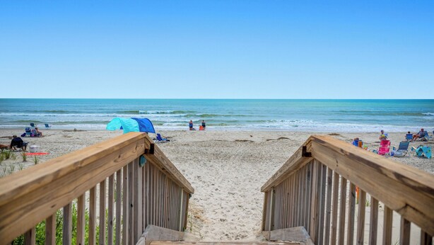 Eine Holzterrasse führt zu einem Strand mit einem Zelt und Strandstühlen.