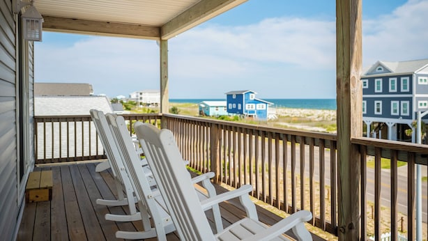 Eine Holzterrasse mit weißen Schaukelstühlen und Blick auf Strand und Meer.