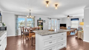 A spacious kitchen with a large island, wooden dining table, and chairs. There is a ceiling fan and pendant lights.