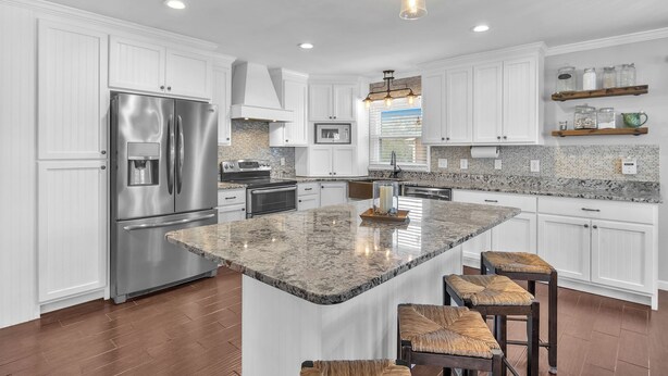 A modern kitchen with white cabinetry, a stainless steel refrigerator, granite countertops, and a central island with bar stools.