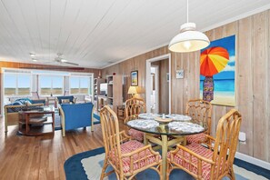 A room with a dining table, wicker chairs, a ceiling fan, and a large window. - Late for the Sky: Ocean and Waterway Views, Near Beach Access (Caswell Beach)